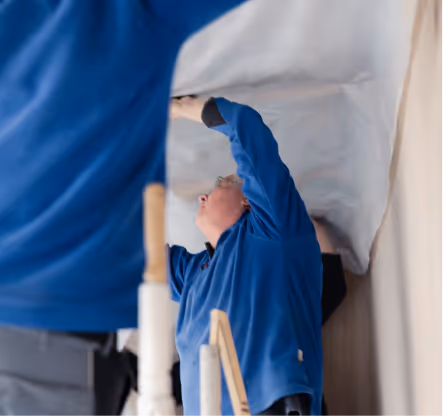 Person in blauem Pullover breitet ein weißes Bettlaken über ein Bett aus.