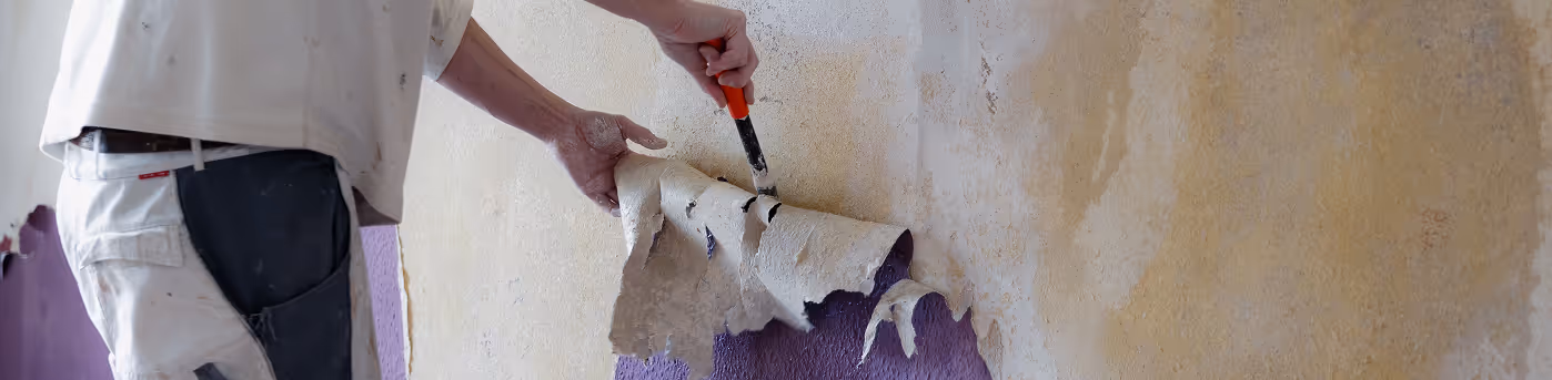 Person entfernt Wandtapete mit einem Spachtel, während darunter eine lila Wand sichtbar wird.