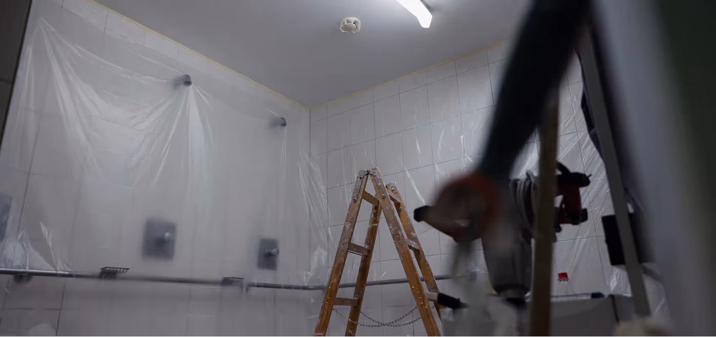 Zimmer mit weißen Fliesenwänden, einer Holzleiter und Plastikfolie, die für Renovierungsarbeiten hängt.